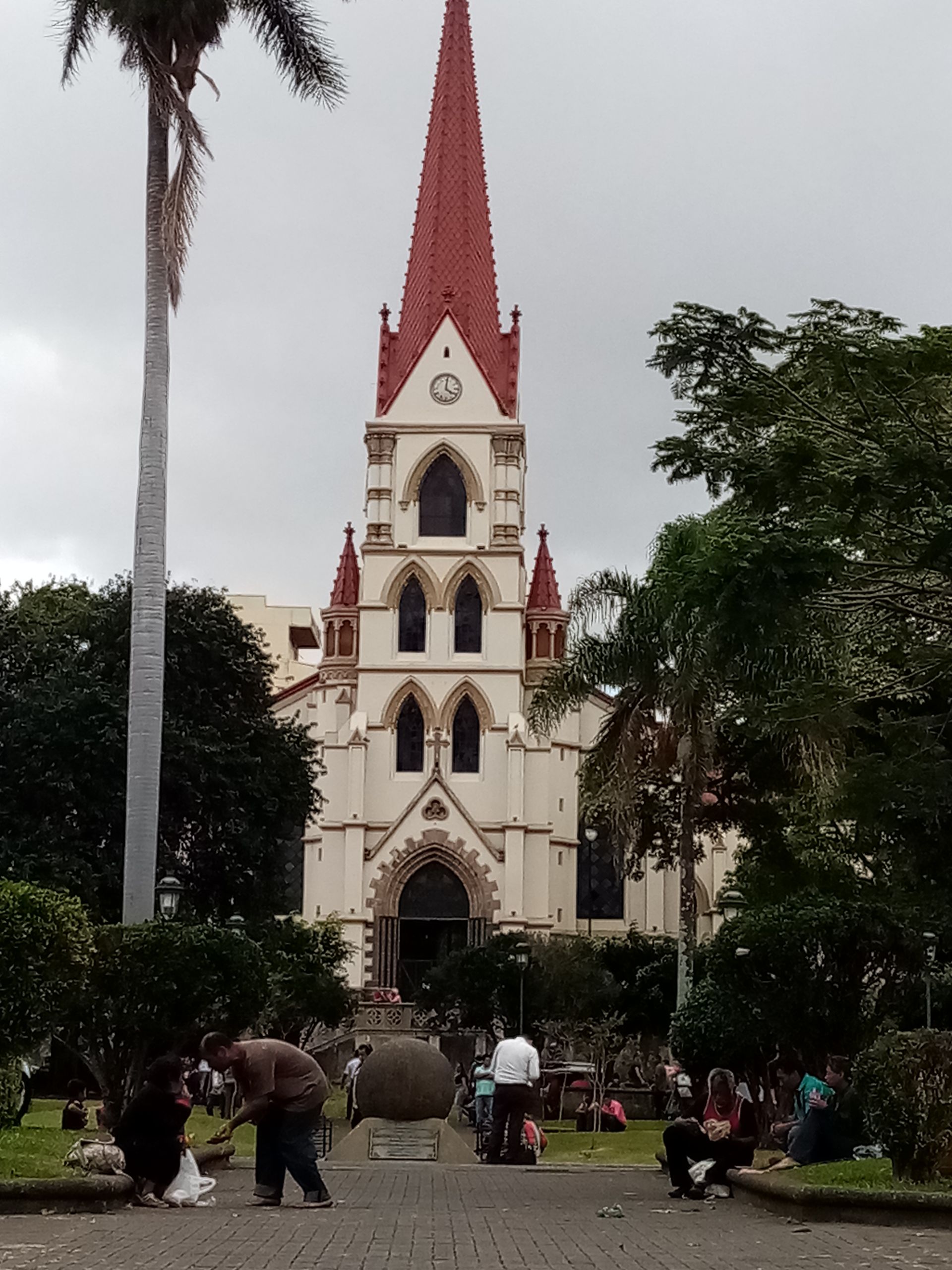 Church with tall red spire against cloudy sky. People gather in front of the building.