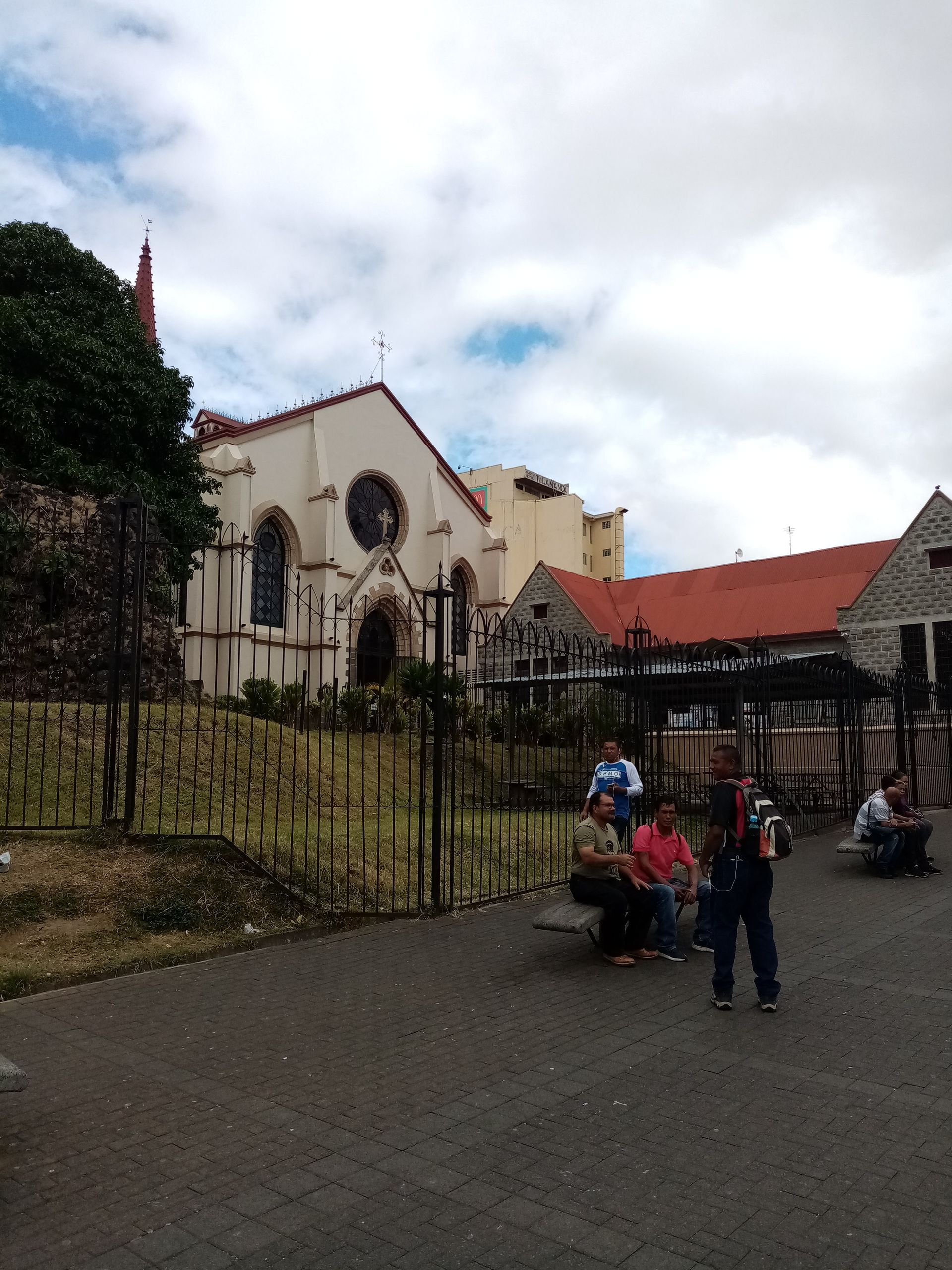 Church building with white walls, red roof, and black fence; people sit and stand near it. Cloudy sky.