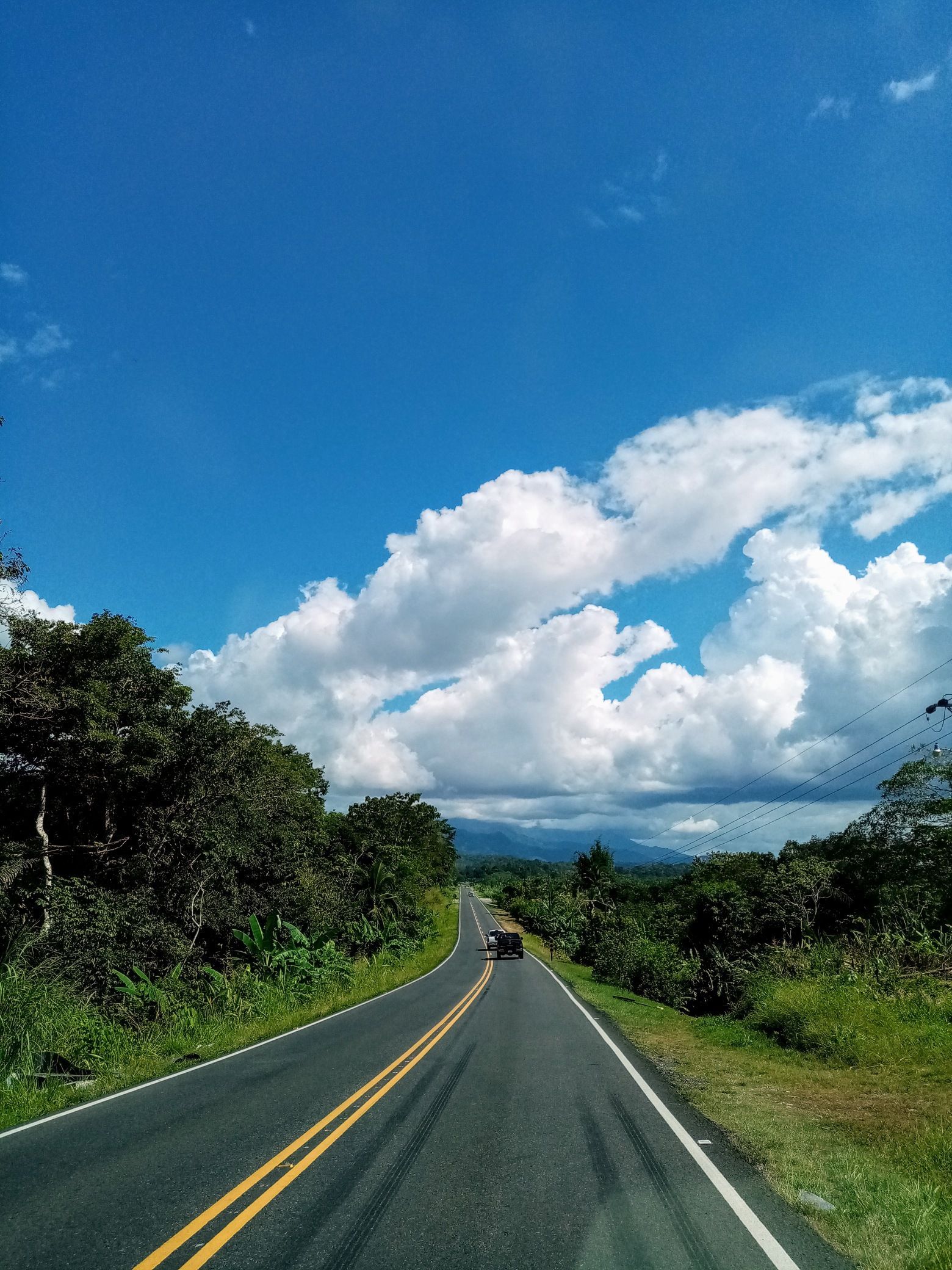 Road through lush greenery under a bright blue sky with puffy white clouds, car driving ahead.
