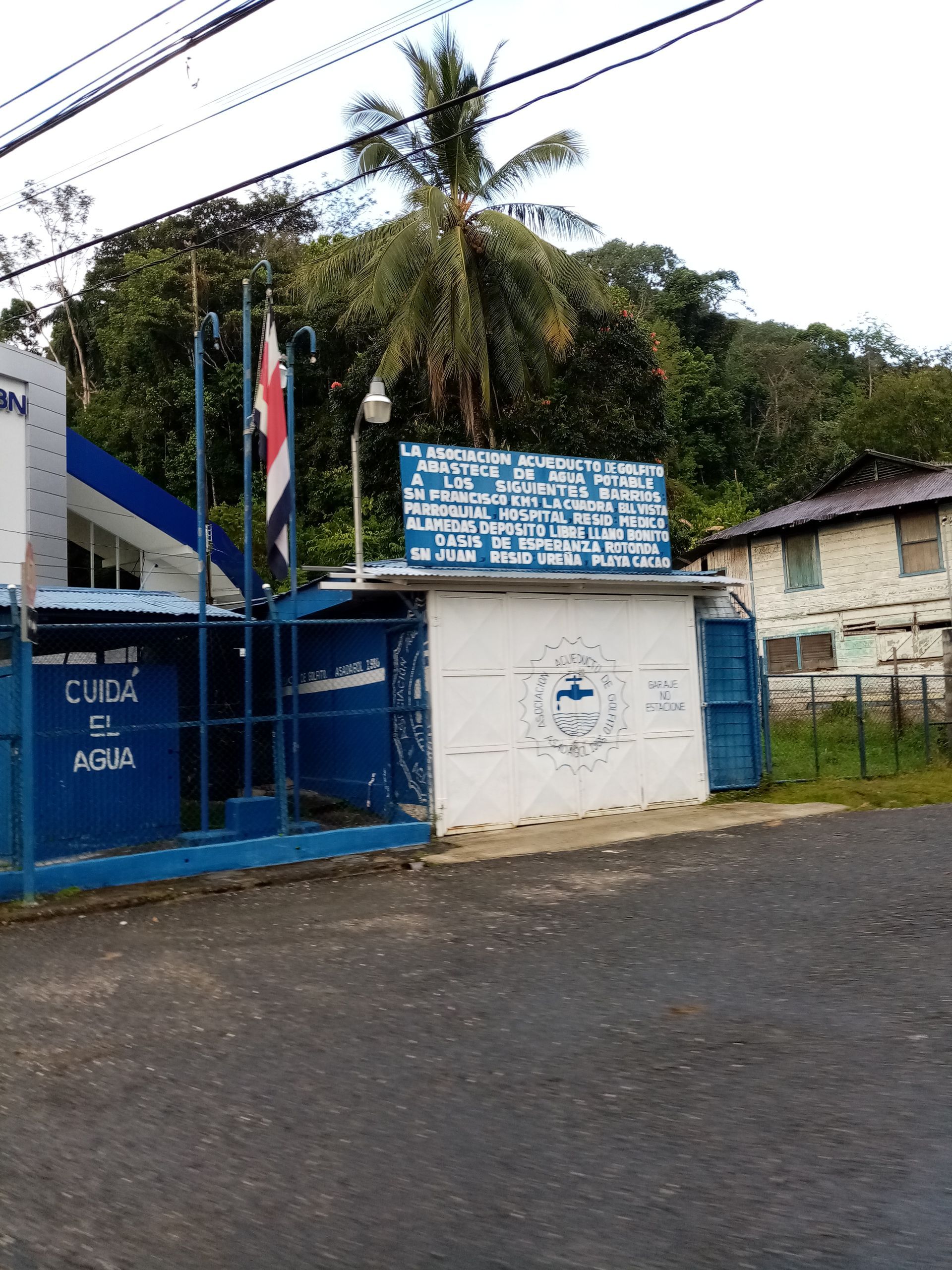 A blue and white building with a Costa Rican flag; a sign above the entrance; trees in background.