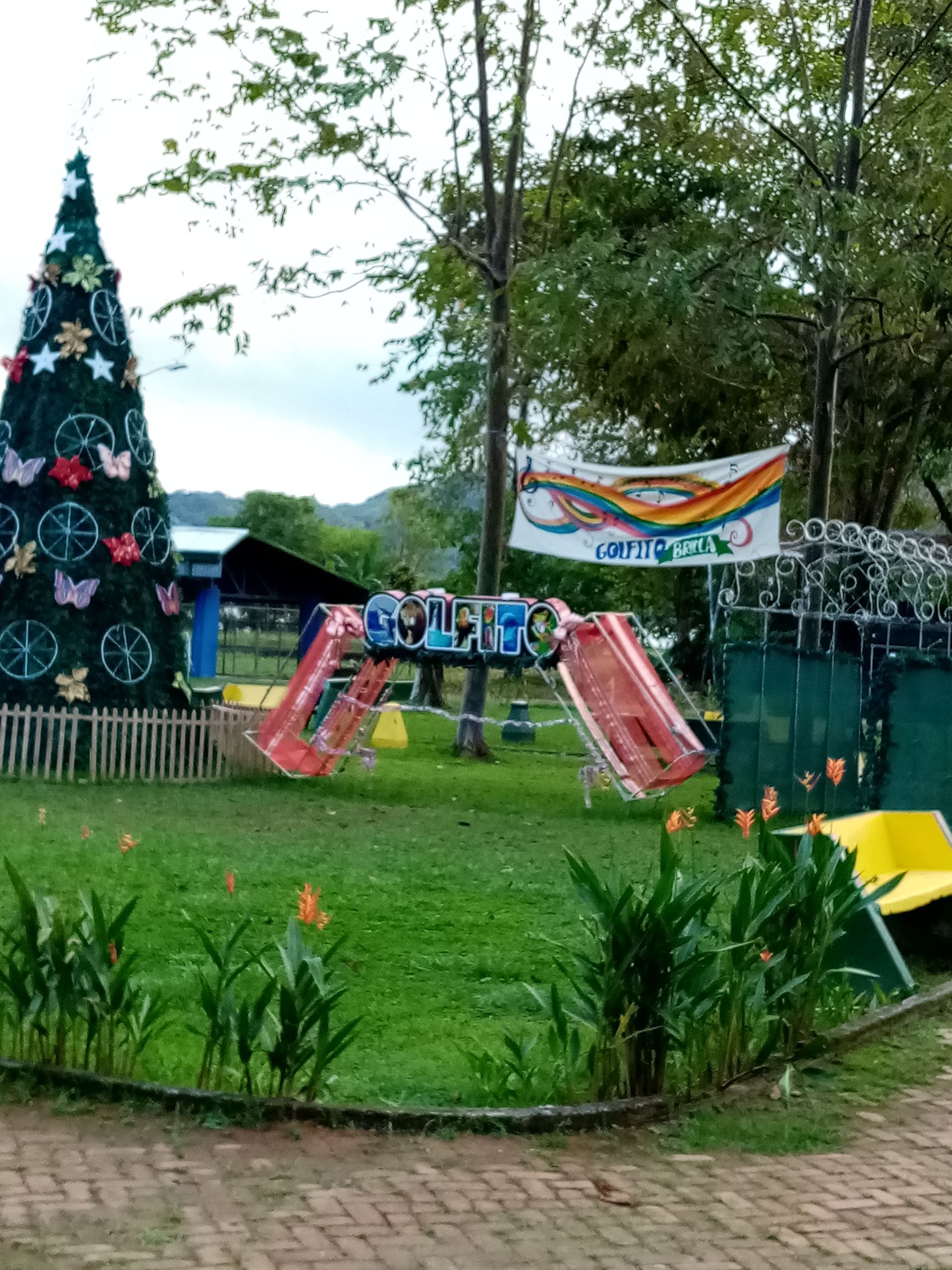 A park scene with a Christmas tree, playground equipment, and greenery. A banner hangs in the background.