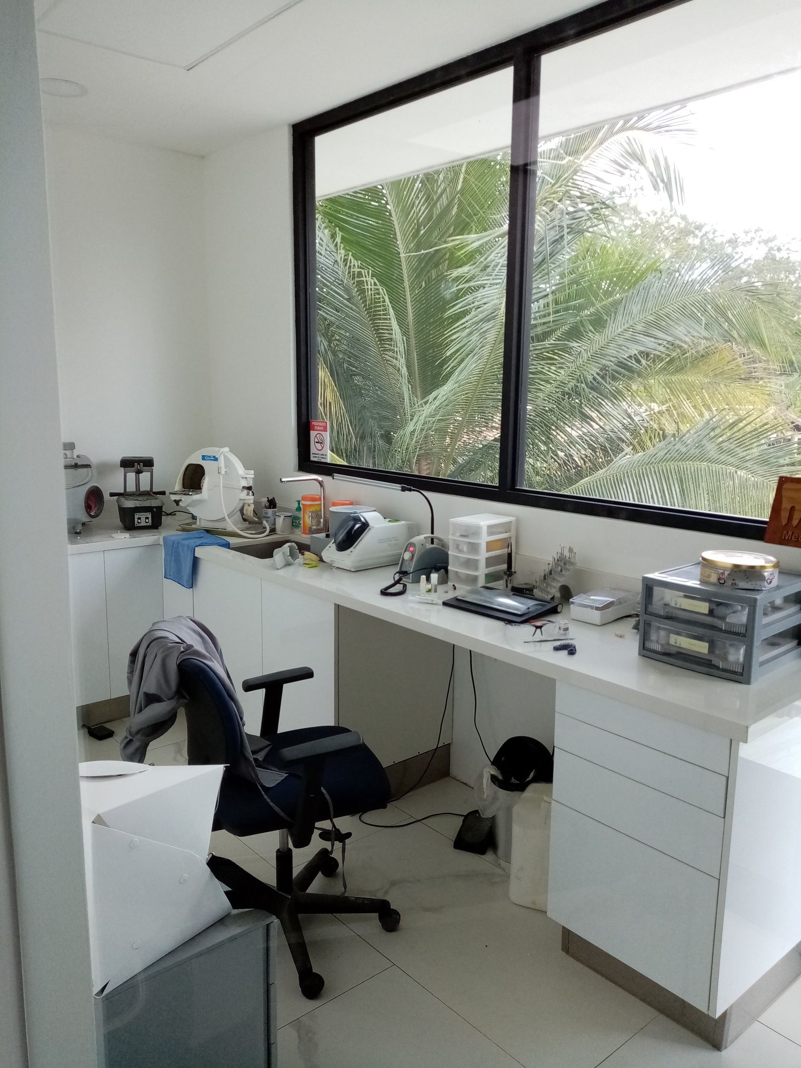 Messy office with desk by a large window overlooking trees. A black office chair sits in front.