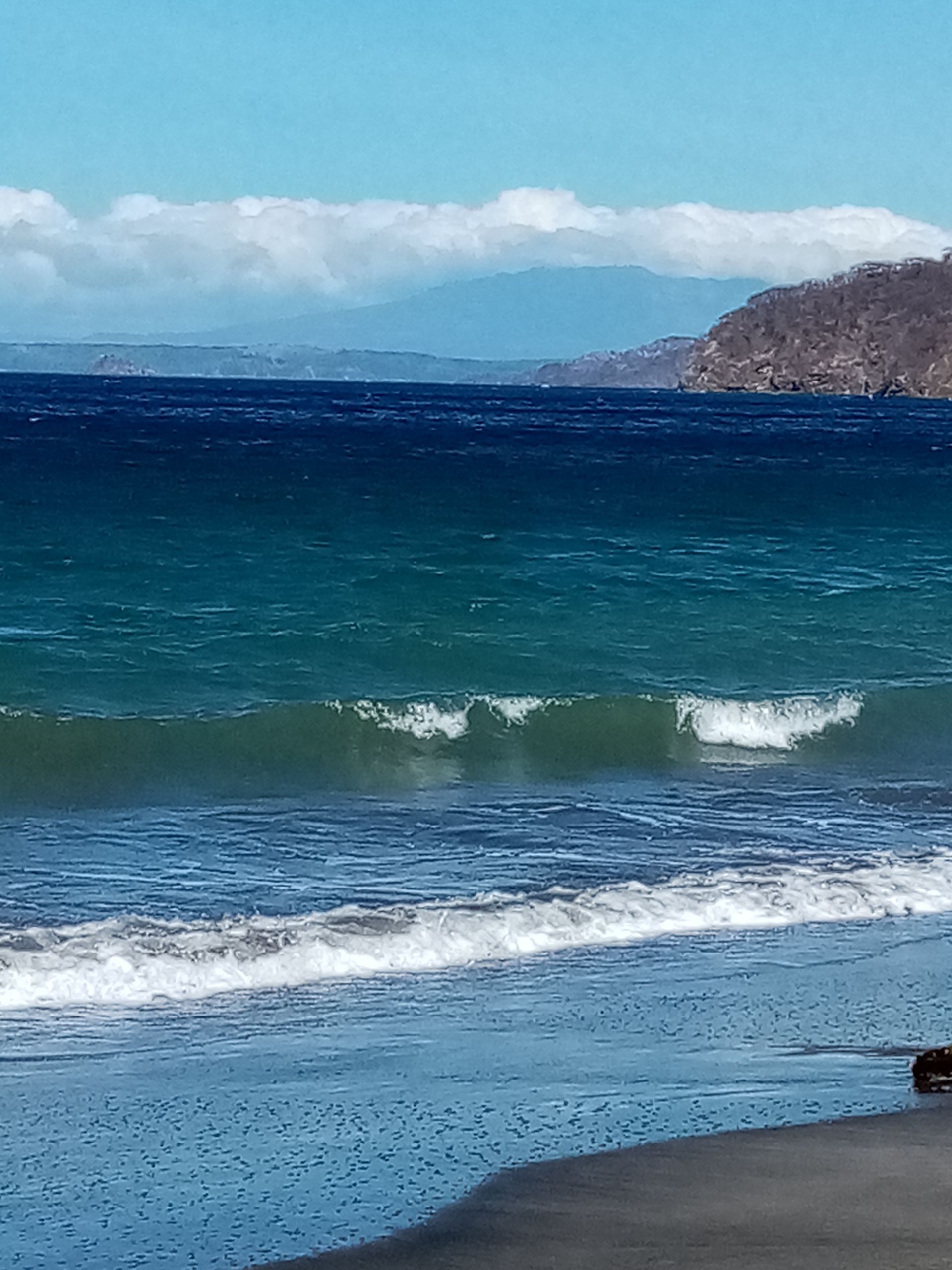 Ocean waves crashing on a dark sandy beach under a blue sky, with mountains in the distance.