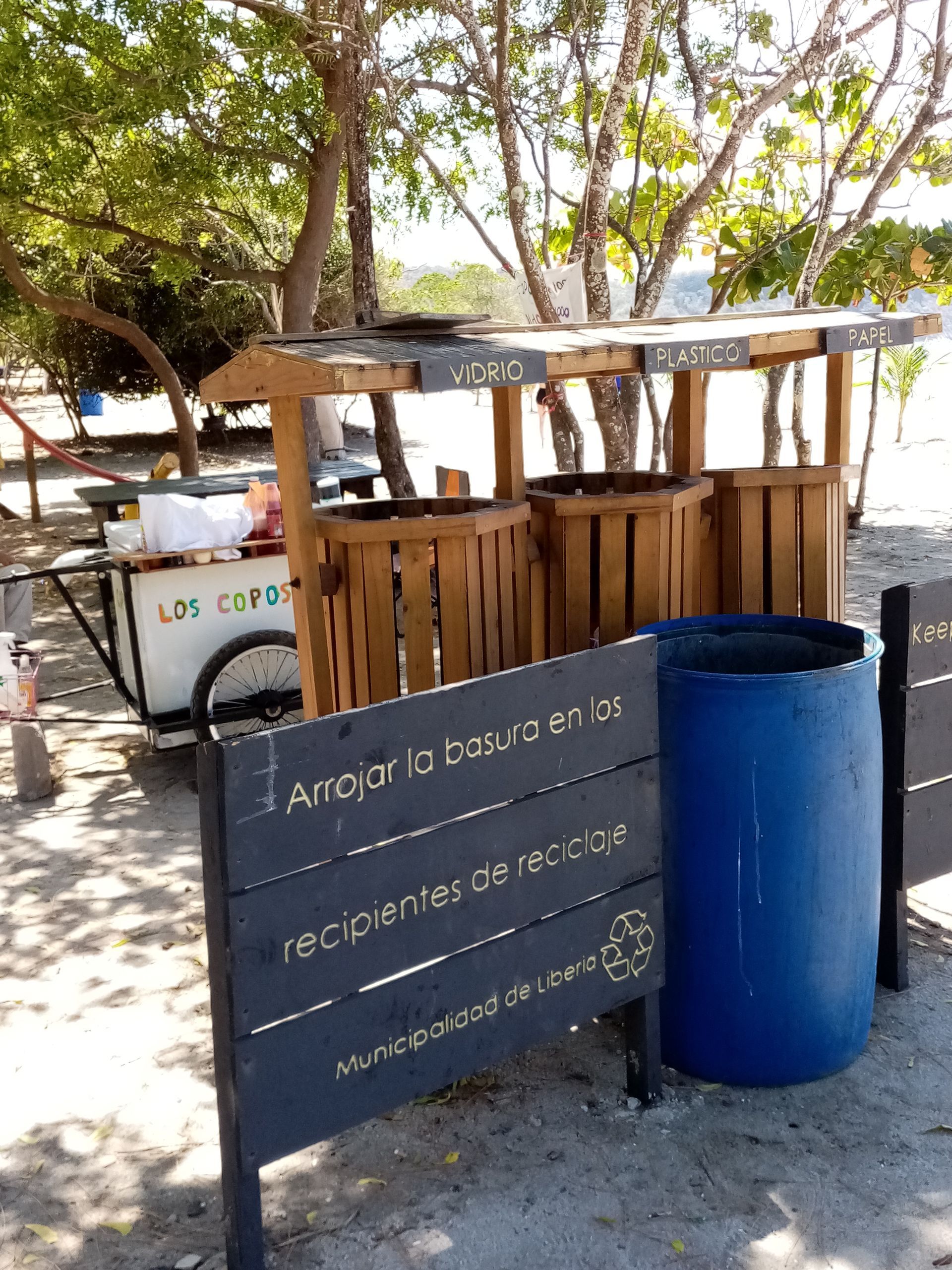 Wooden recycling bins on a beach. A sign urges proper disposal. A vendor cart is nearby.