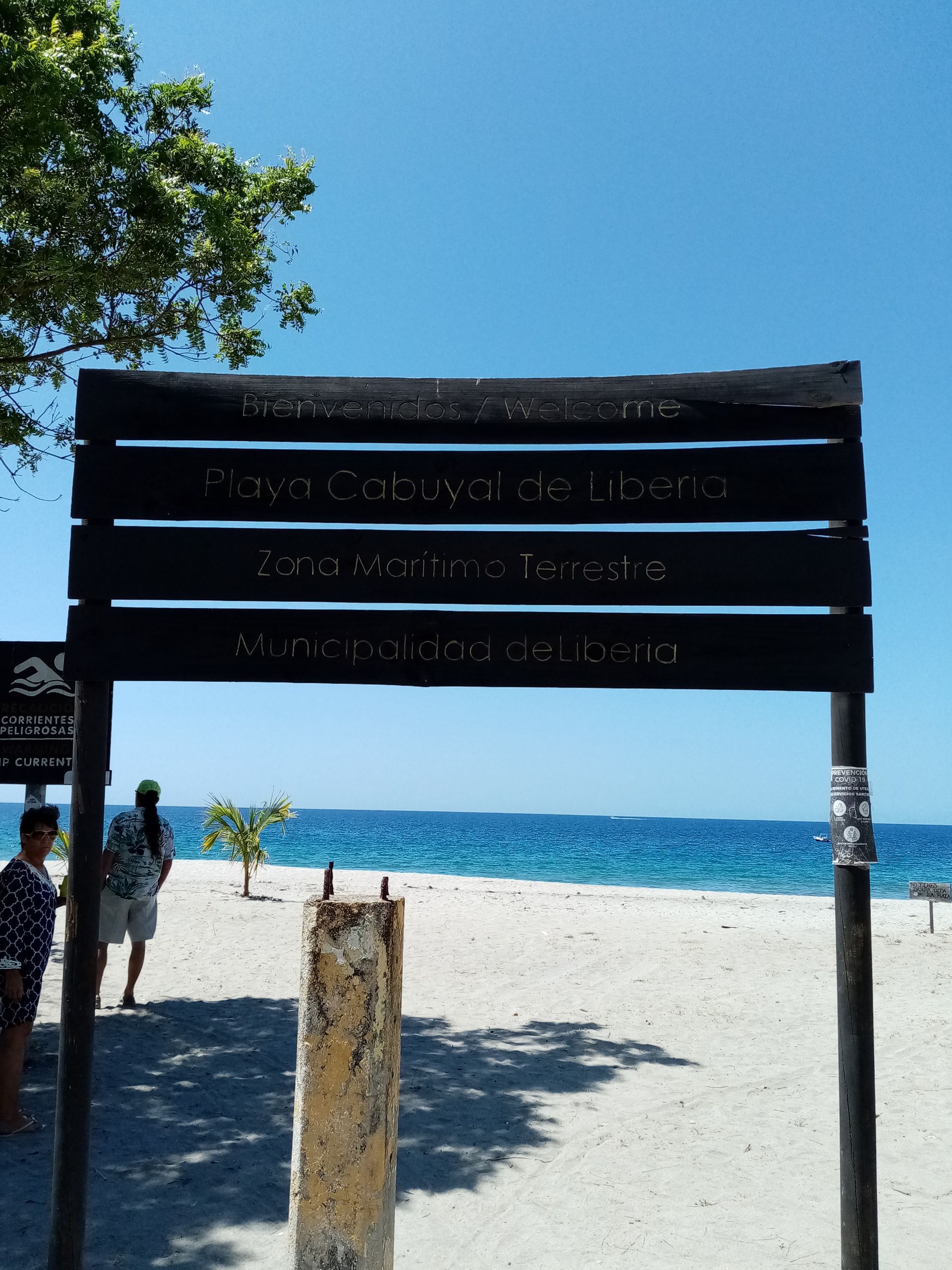 Sign at a beach with text; blue ocean and sky in background; people present.