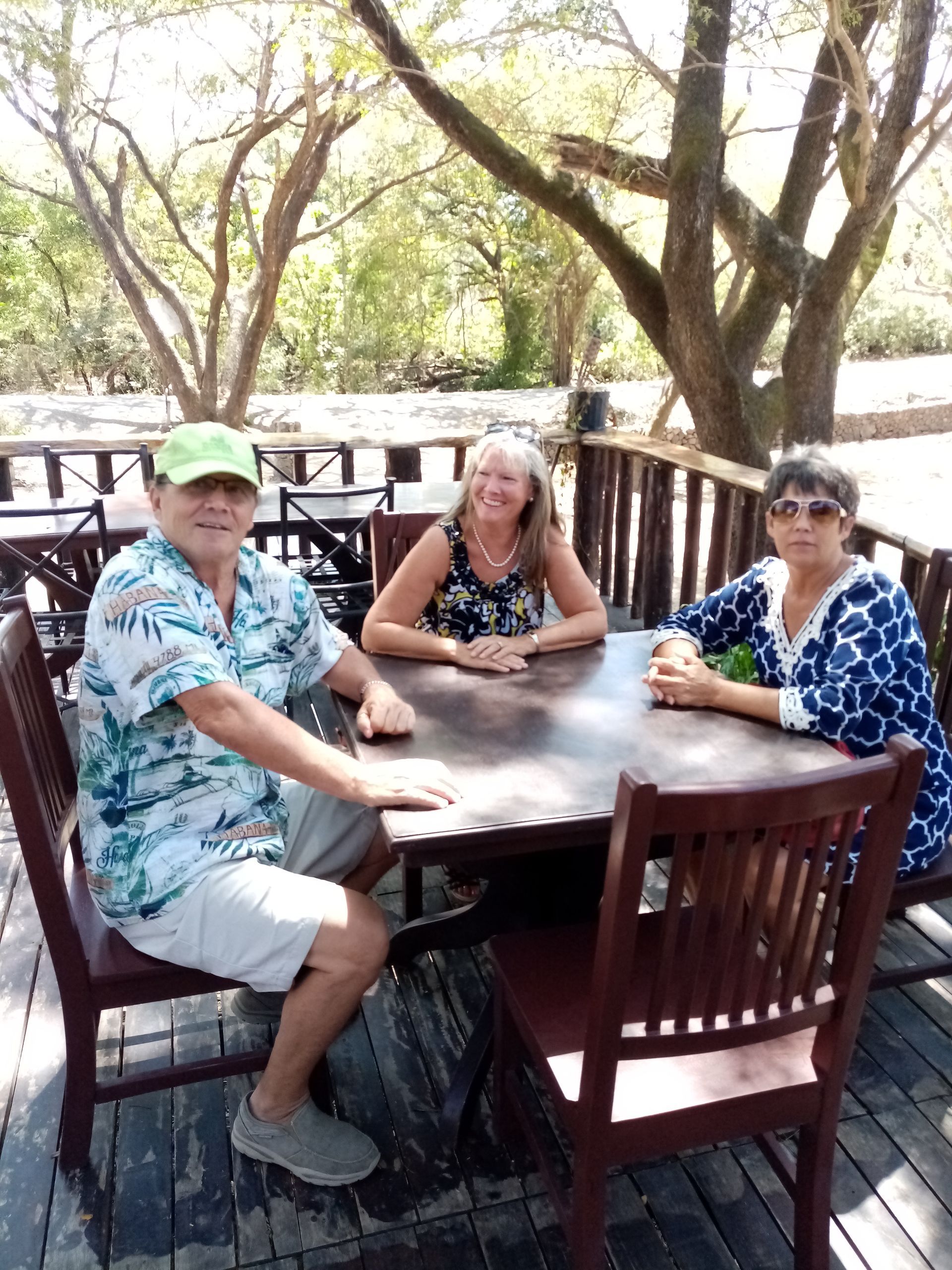 Three people seated at a table on a wooden deck outdoors, trees in background.