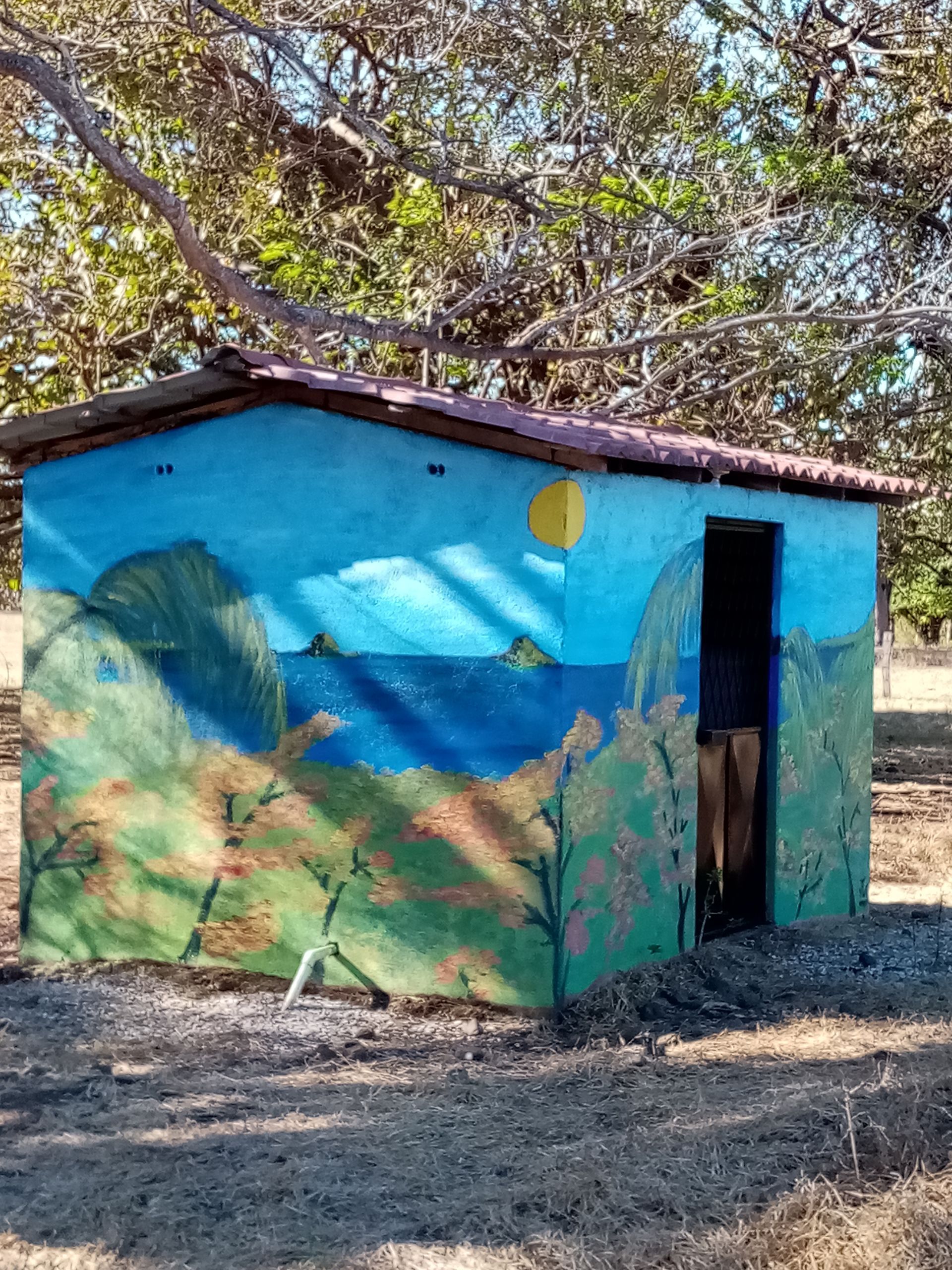 Small, concrete building with painted landscape scene; blue sky, sun, ocean, trees. Brown roof and open doorway.