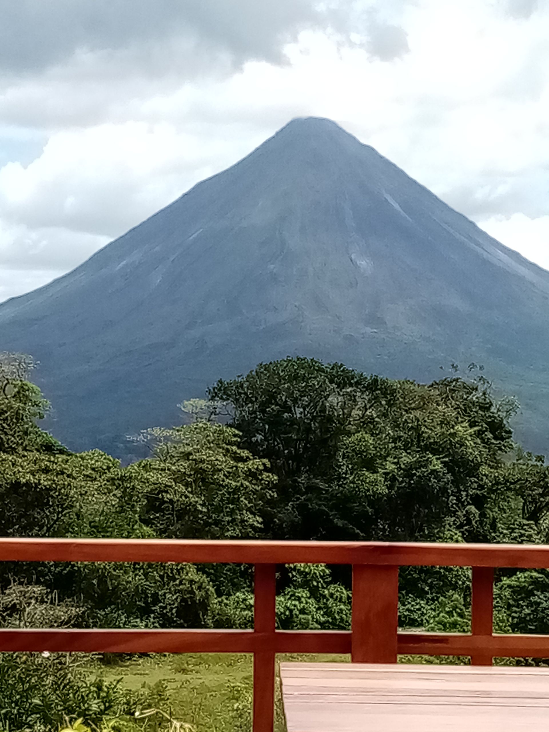 Volcanic cone rises above lush green treetops, viewed from a wooden deck under a cloudy sky.