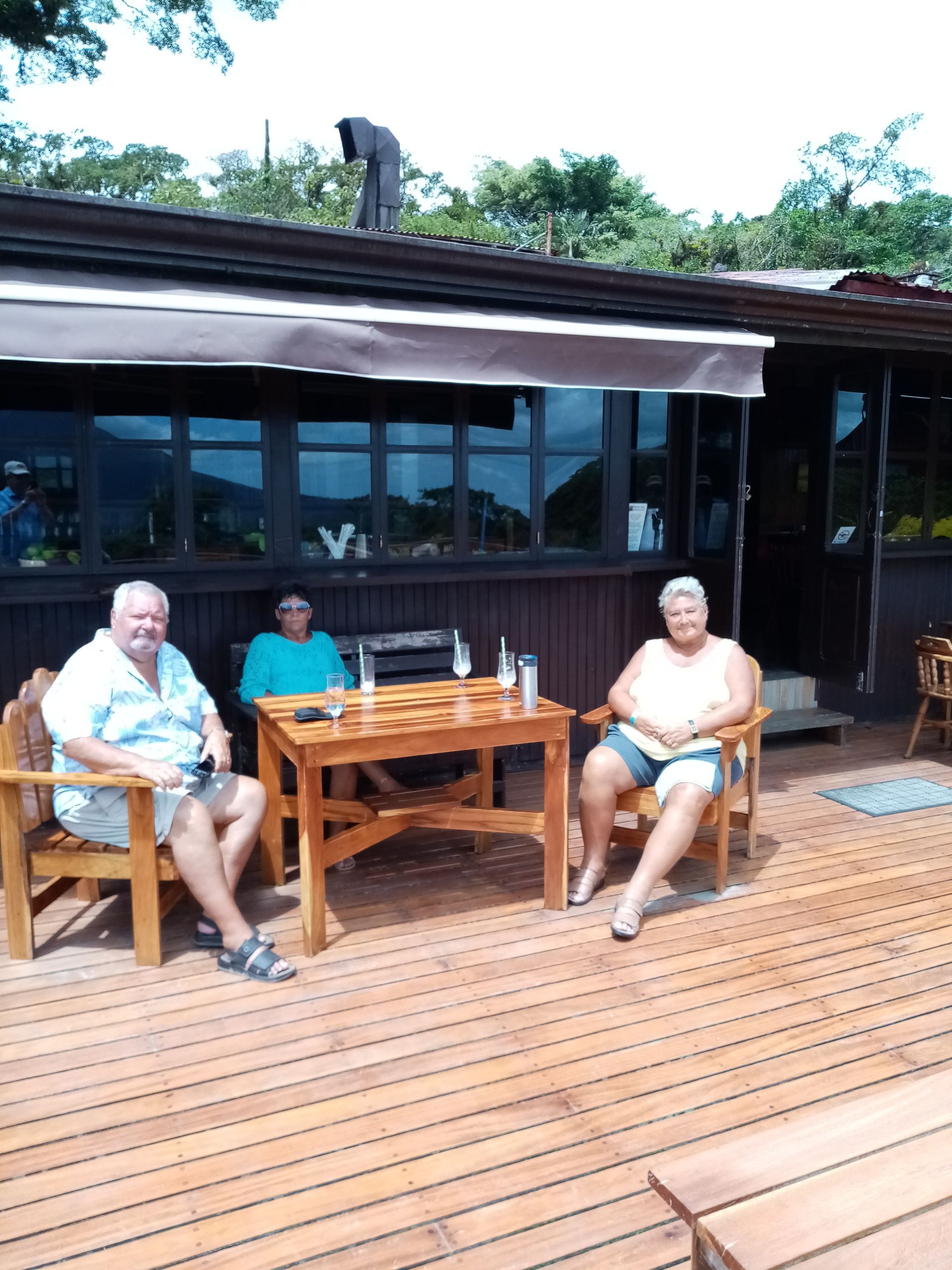 Three people seated at a wooden table on a deck; building with windows in background.
