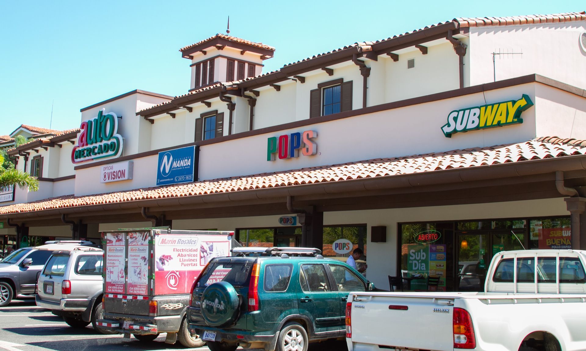 A strip mall with a Subway and other shops; cars are parked in front.