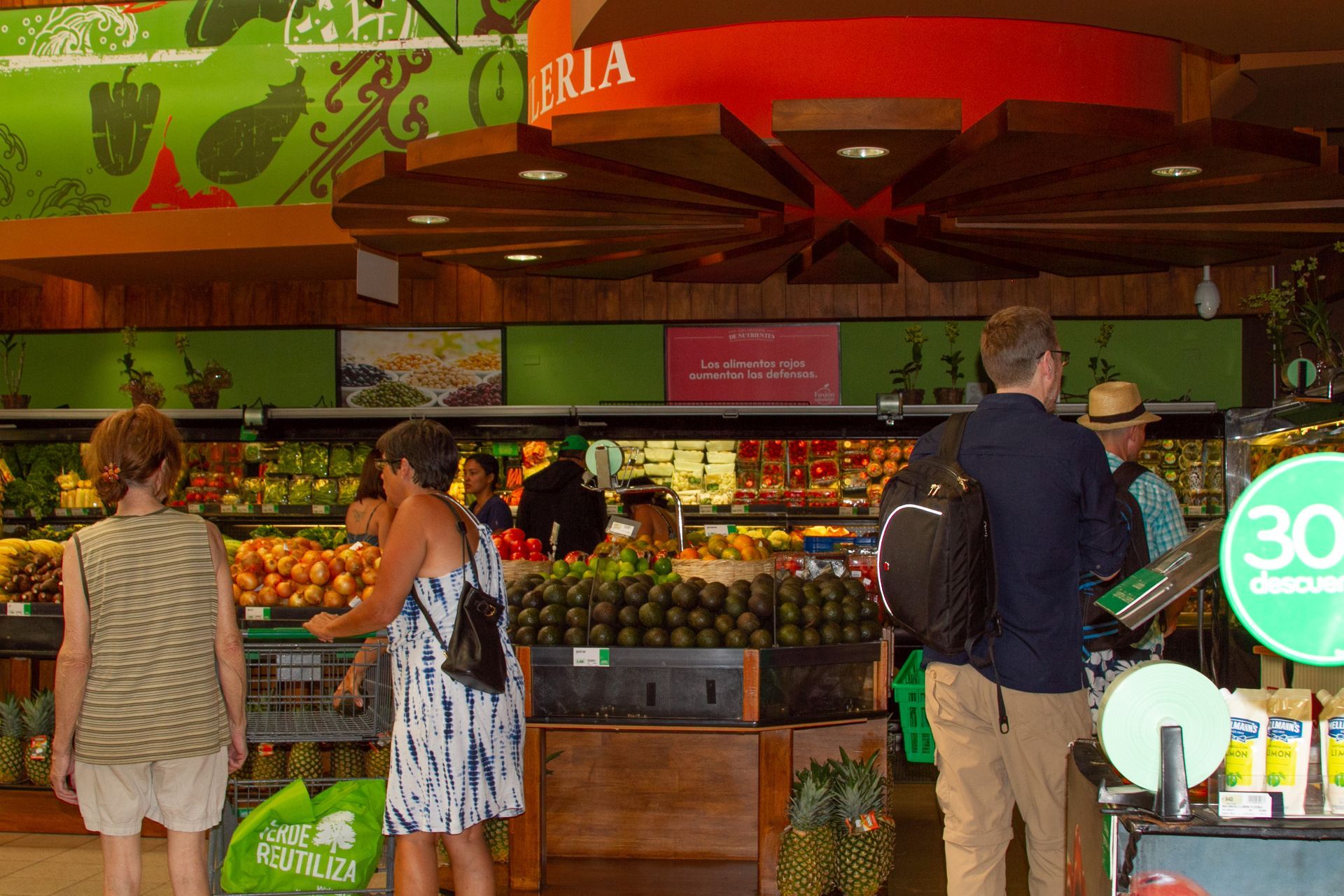 People shopping for produce in a brightly lit grocery store. Displays include fruit, overhead signage, and shoppers.