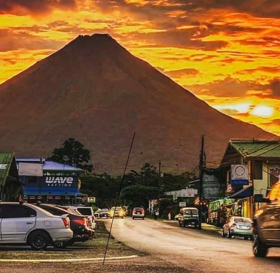 Cars parked along road in front of village, volcano at sunset with orange sky.