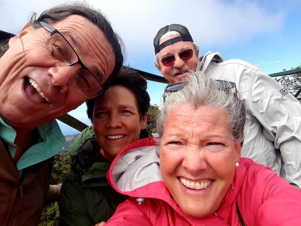 Four people smiling, taking a selfie outdoors, blue sky visible.