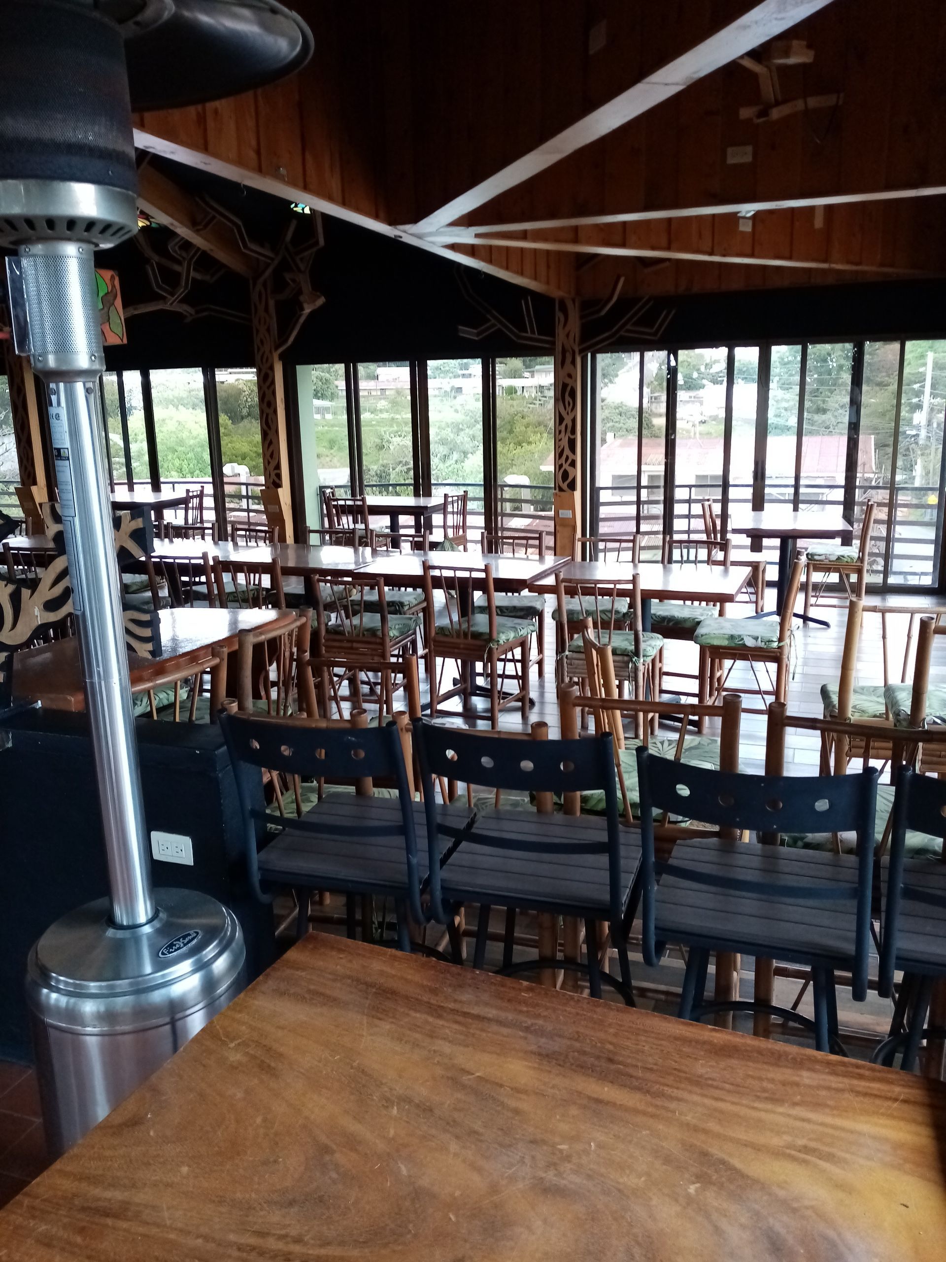 Restaurant interior with wooden tables and chairs, large windows, and a silver patio heater.
