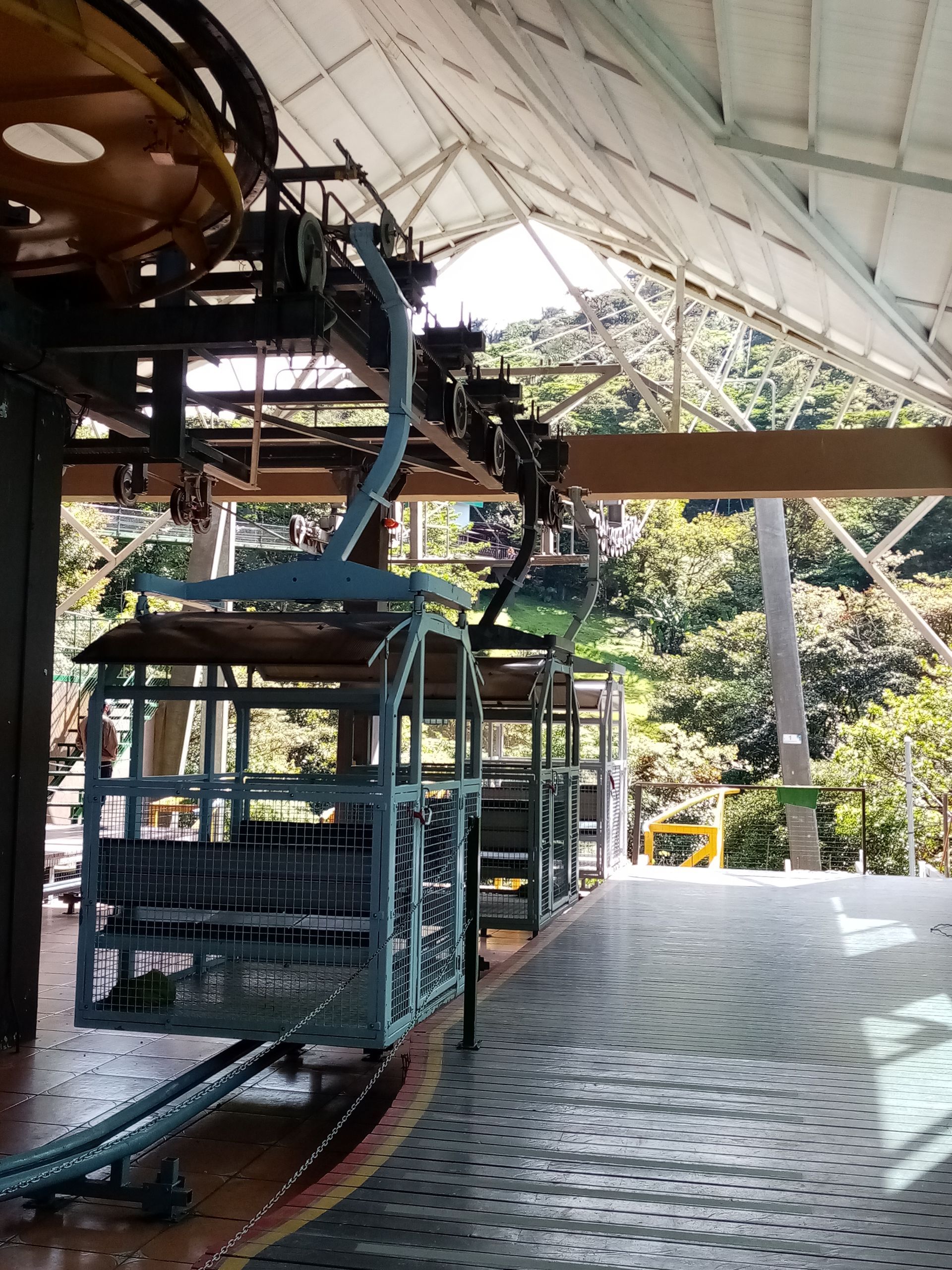 Cable car station, blue and yellow cars in a row, under a white roof. Lush green trees in the background.
