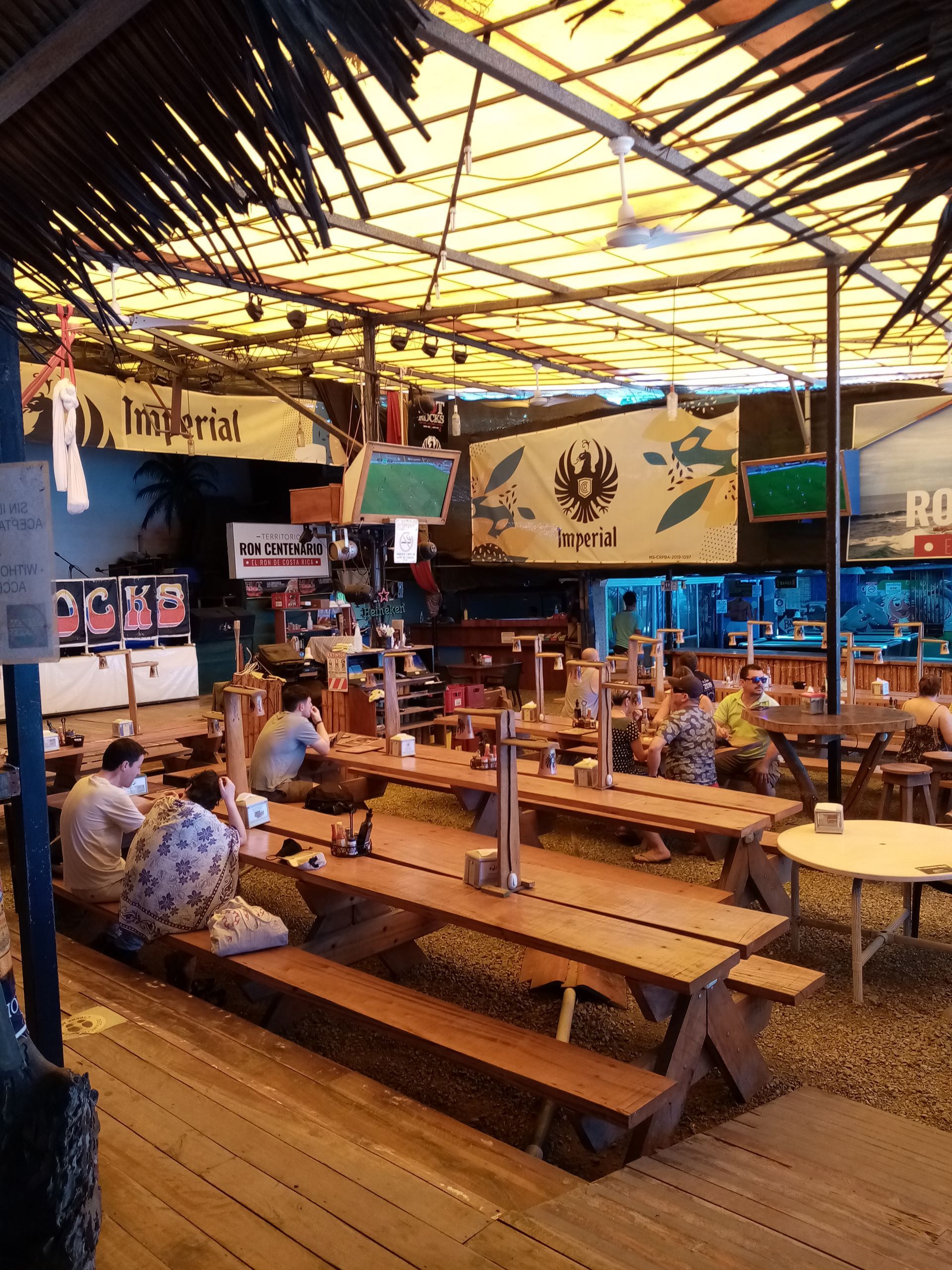 Outdoor restaurant with wooden tables and benches, people seated, under a yellow canopy.