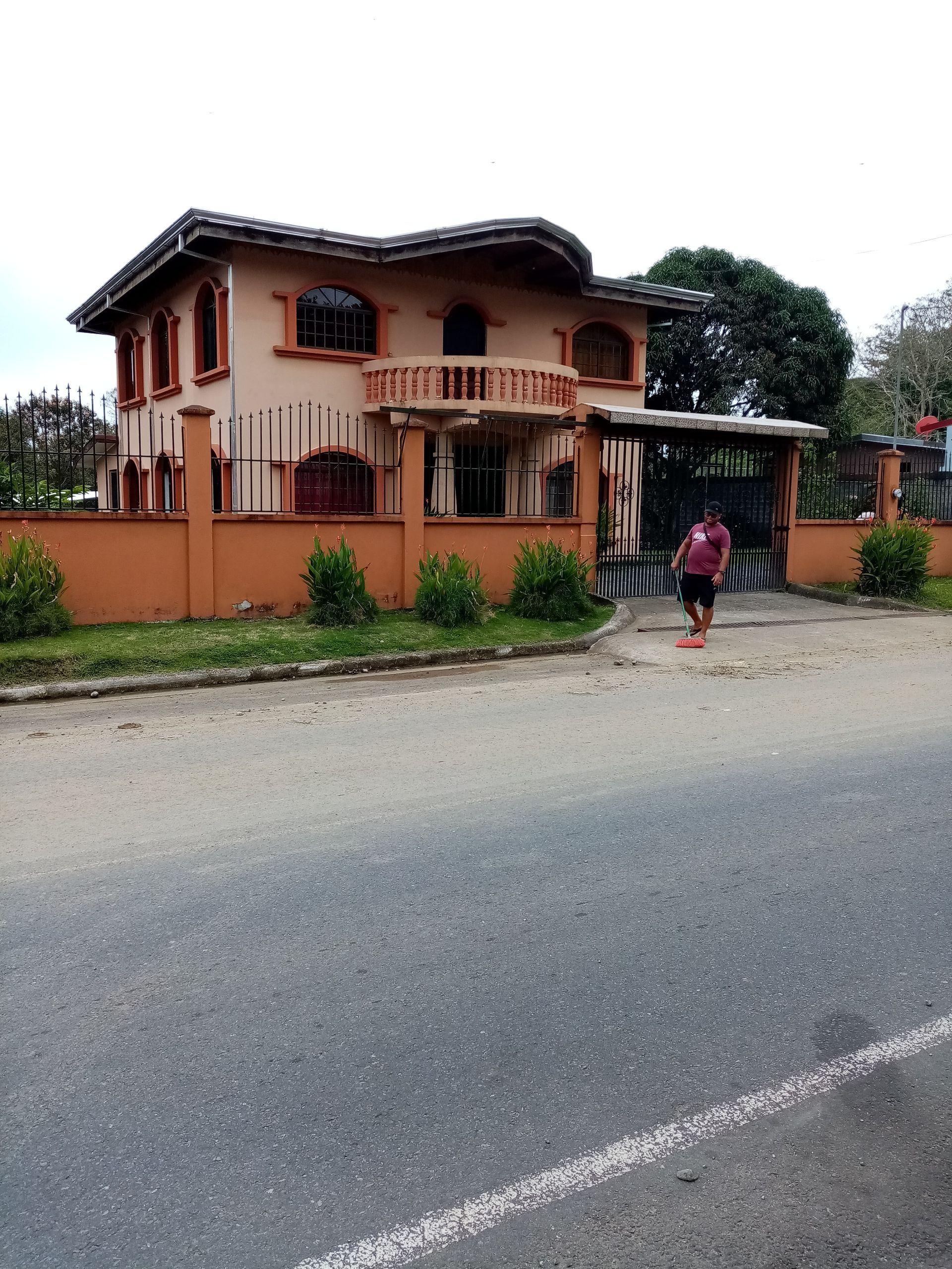 Two-story beige house behind an orange wall, person walking near the gate, road in the foreground.