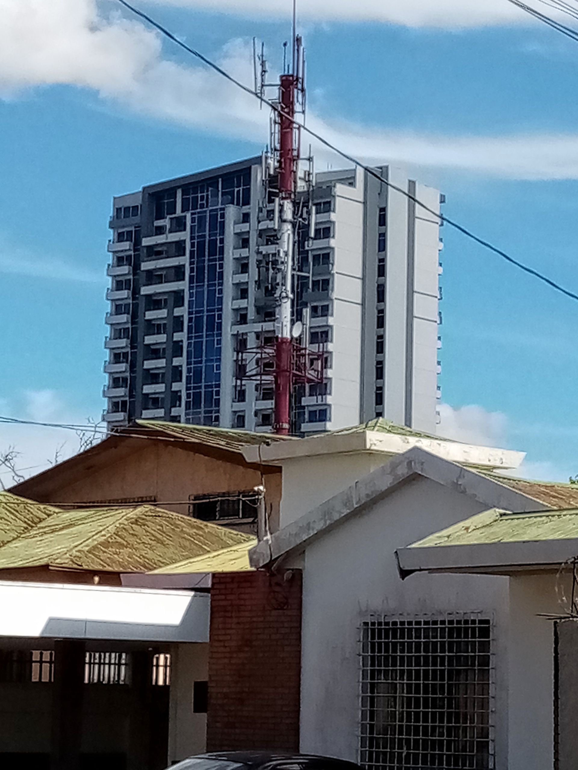 Tall building with antenna on top, seen above rooftops, against a cloudy blue sky.