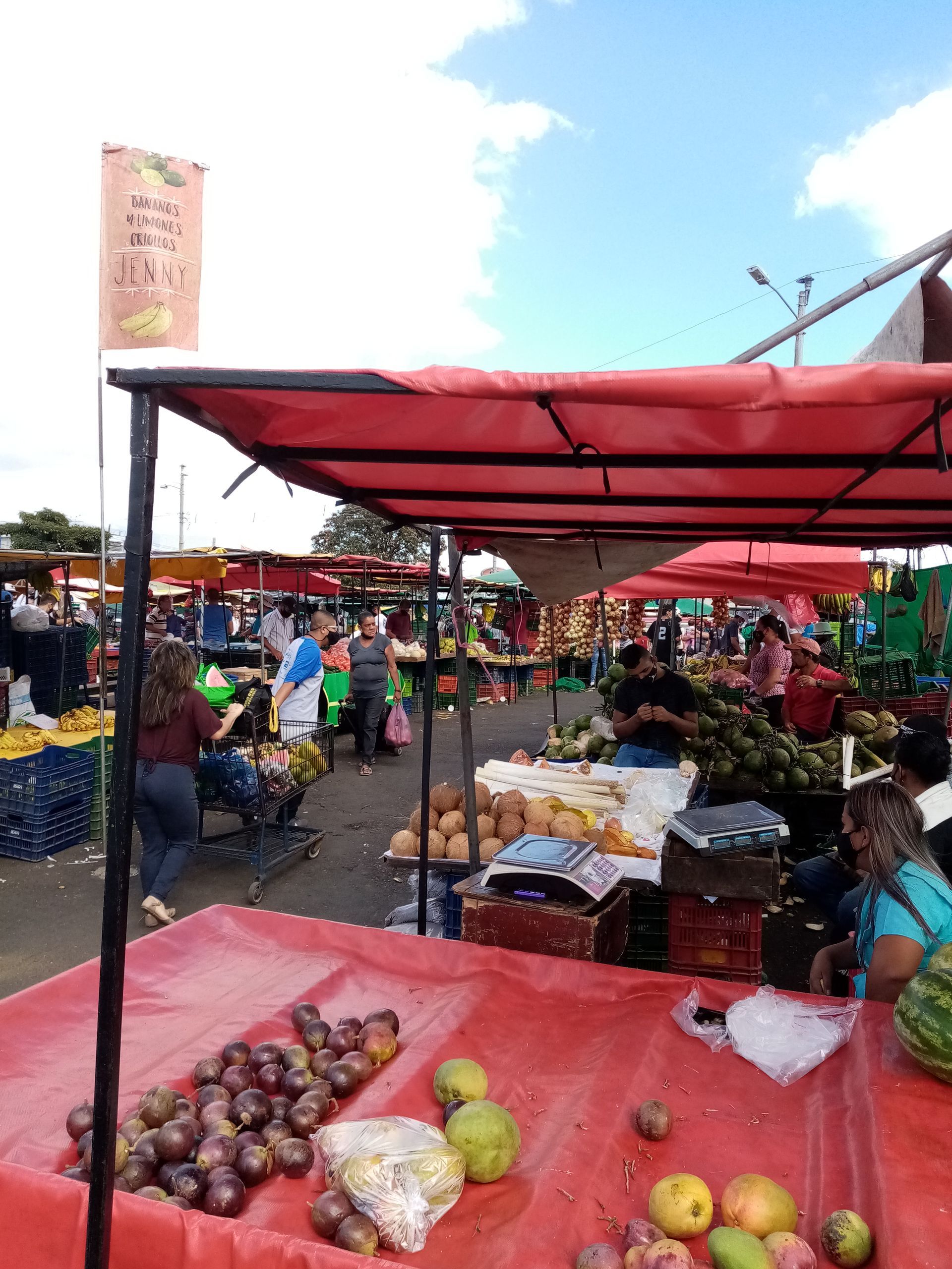 A bustling outdoor market with red-covered stalls selling fruits and vegetables under a blue sky. People browse.