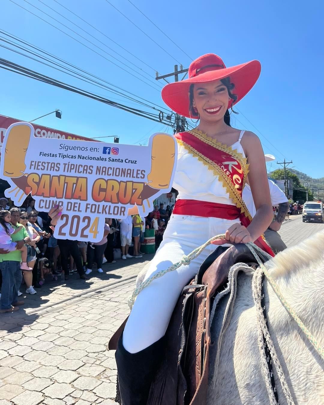 Woman in white and red outfit on horseback, holding banner advertising a festival in Santa Cruz, Costa Rica.