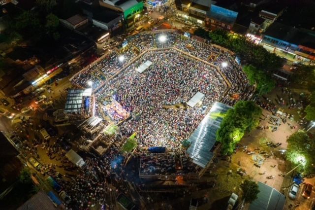 Aerial view of a large nighttime crowd gathered in a city square, lit by stage lights.
