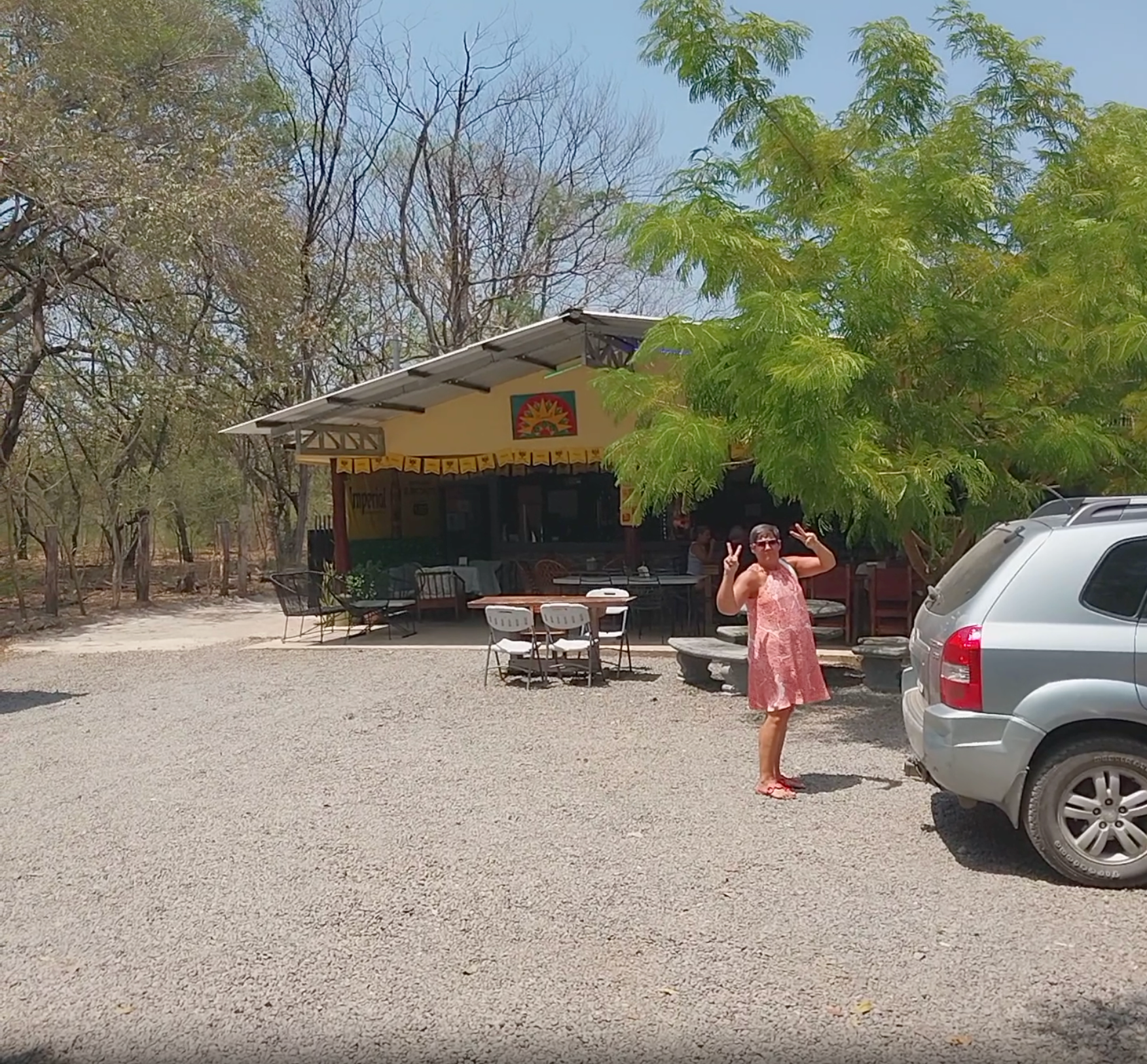 Woman standing outside a yellow building with outdoor seating. A silver SUV is parked nearby on a gravel lot.