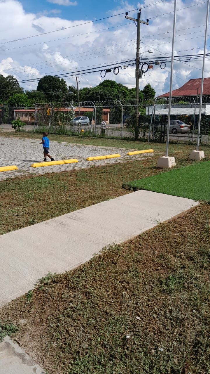 A person walks on a gravel area, beside a sidewalk and grass. Sunny outdoor setting.