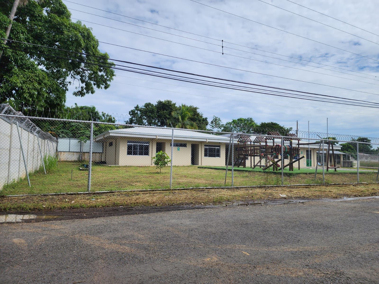 Tan building with several windows, behind a chain link fence and a cloudy sky.