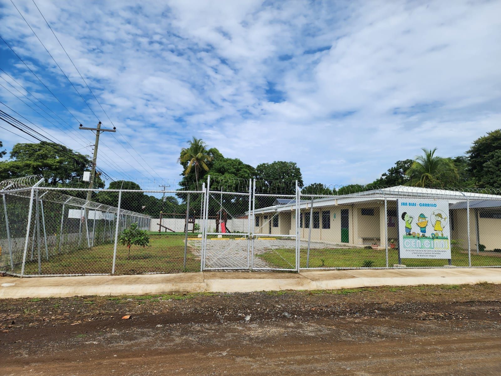 A low, single-story building behind a chain link fence. A sign with children is visible.