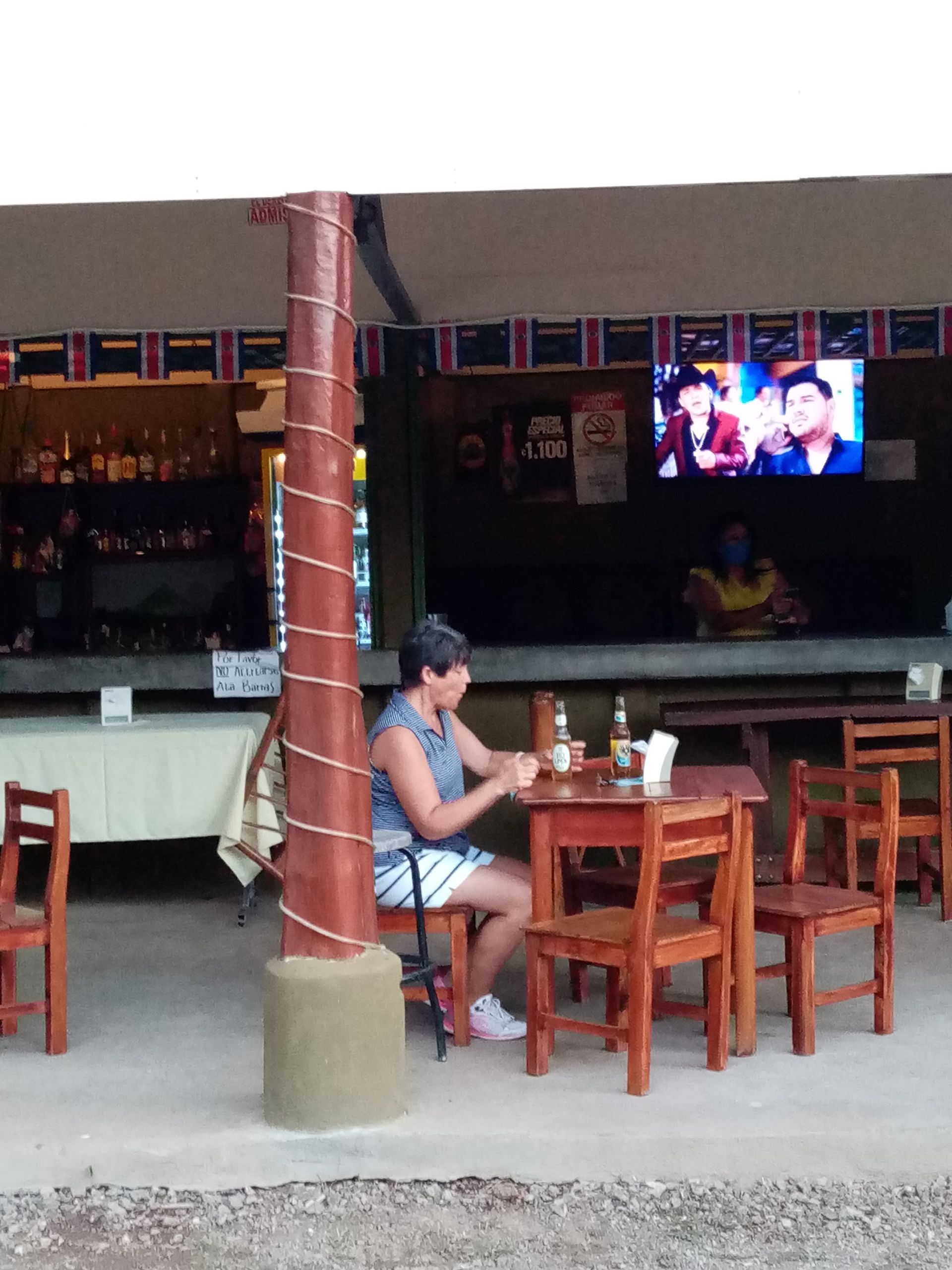 Woman sitting at a small wooden table outside a bar, beer bottles present. Bar has shelves of liquor, TV screen visible.