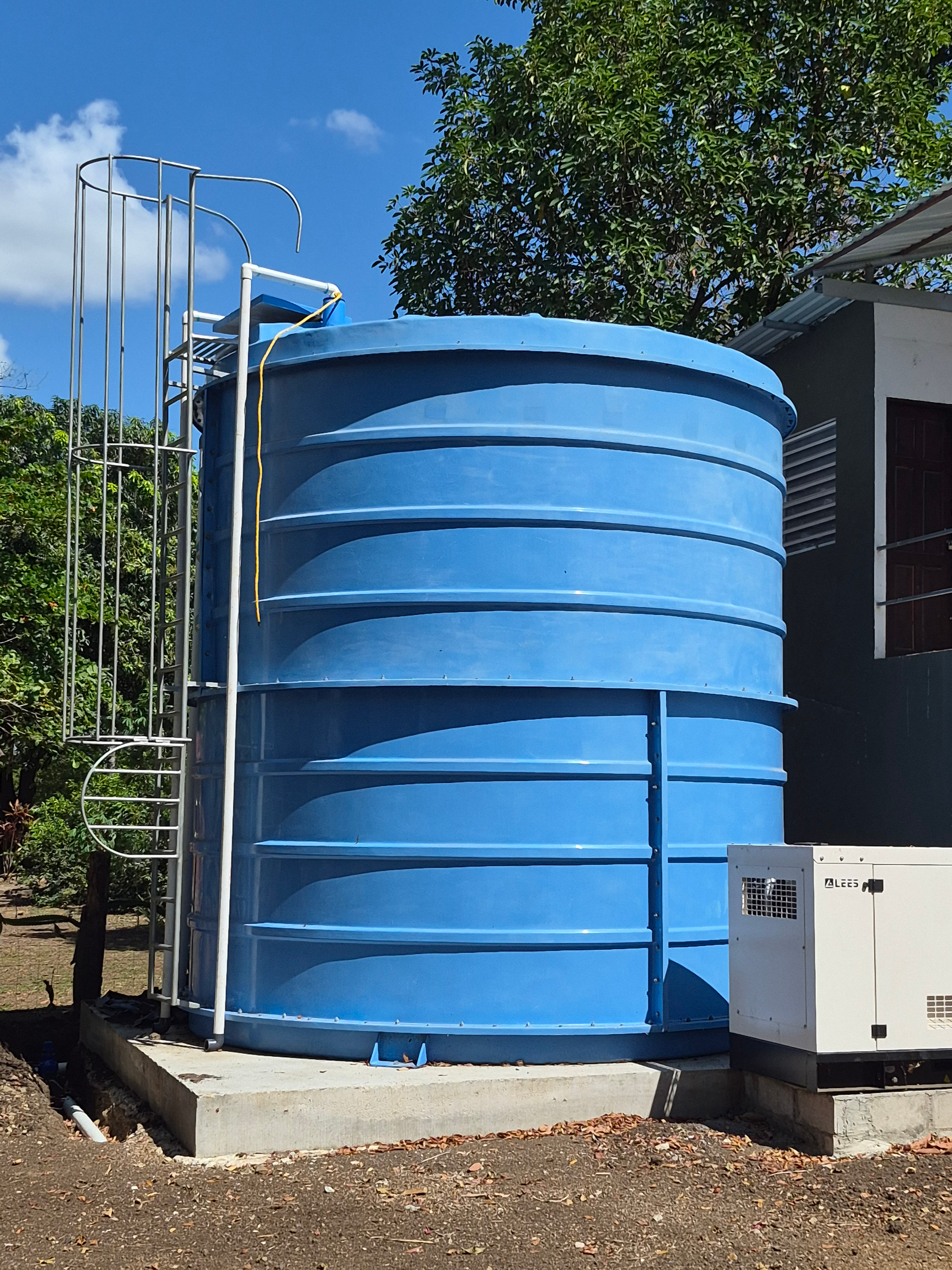 Black water tank on wooden platform, surrounded by green trees in a sunny outdoor setting.