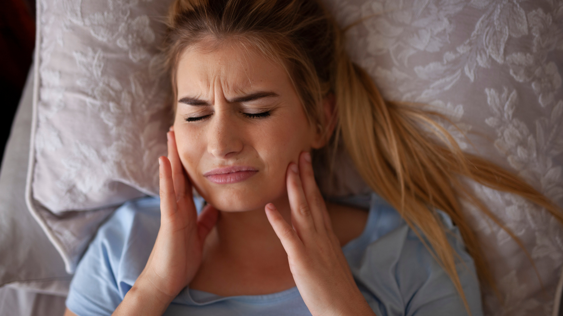 Woman in bed, holding cheeks, facial expression showing pain.
