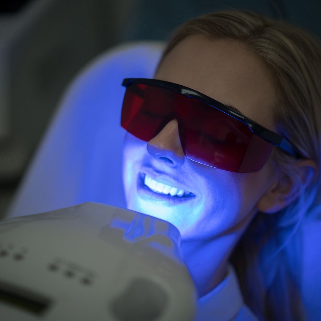 Woman in dental chair with blue light teeth whitening treatment, wearing red protective glasses.