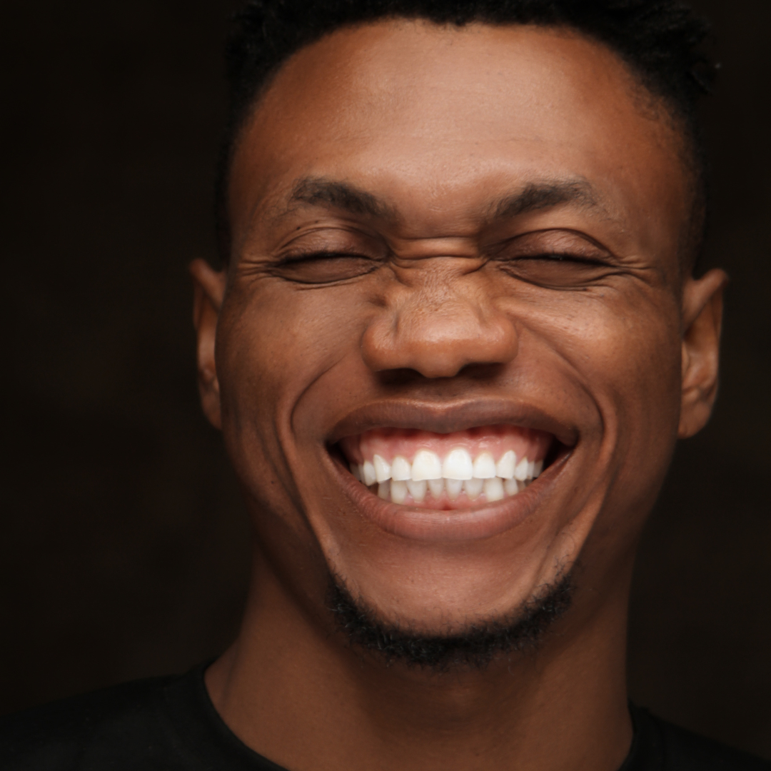 Man with closed eyes, smiling widely, revealing teeth. Dark background.