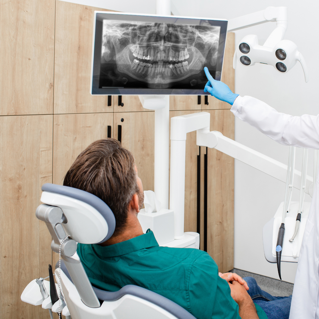 Dentist pointing at patient’s dental X-ray on screen. Patient in chair, looking at the screen. Medical setting.