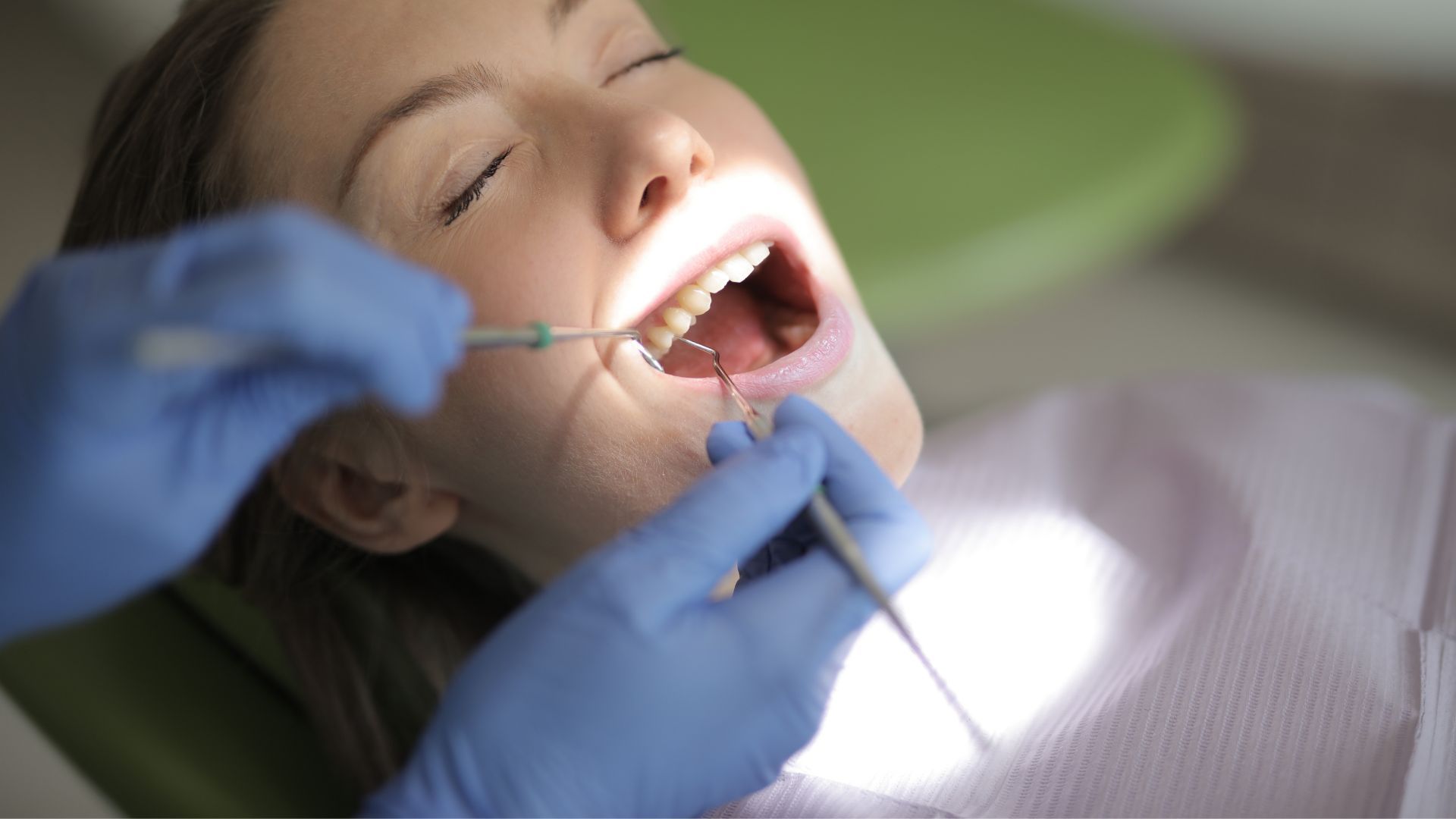 Dentist wearing blue gloves examines a patient’s open mouth with dental tools.