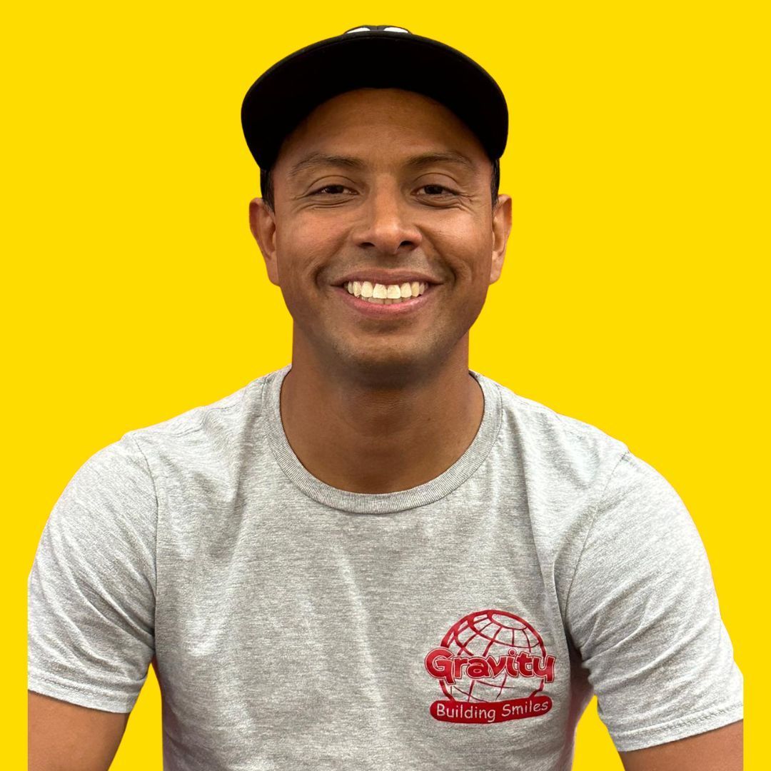 Smiling person in a black cap and gray T-shirt against a bright yellow background.