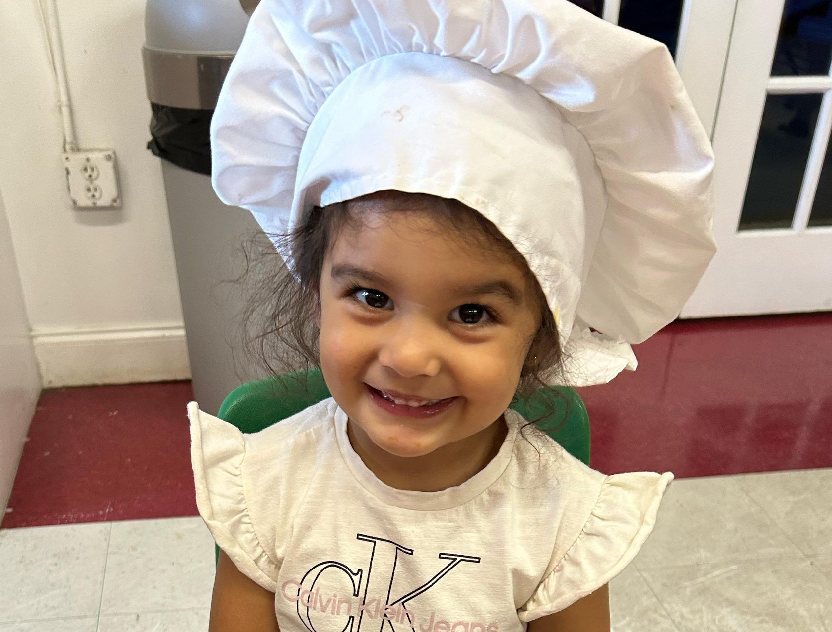 Smiling child wearing a white chef hat and white shirt in a kitchen-like setting