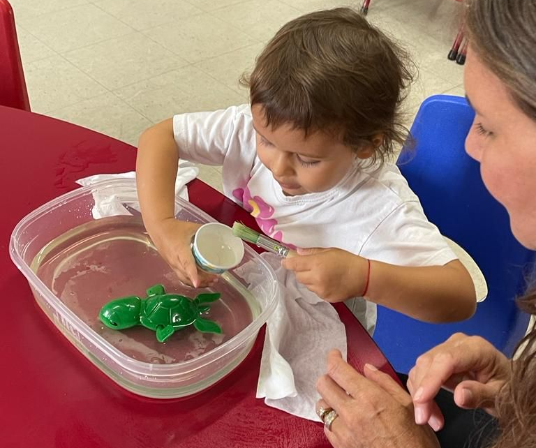 Child scooping green jelly from a clear tub into a bowl at a table with an adult nearby