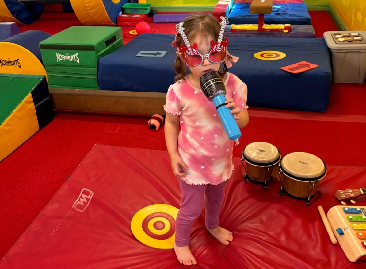 Child in a playroom singing into a toy microphone, with bongo drums and colorful foam mats nearby.