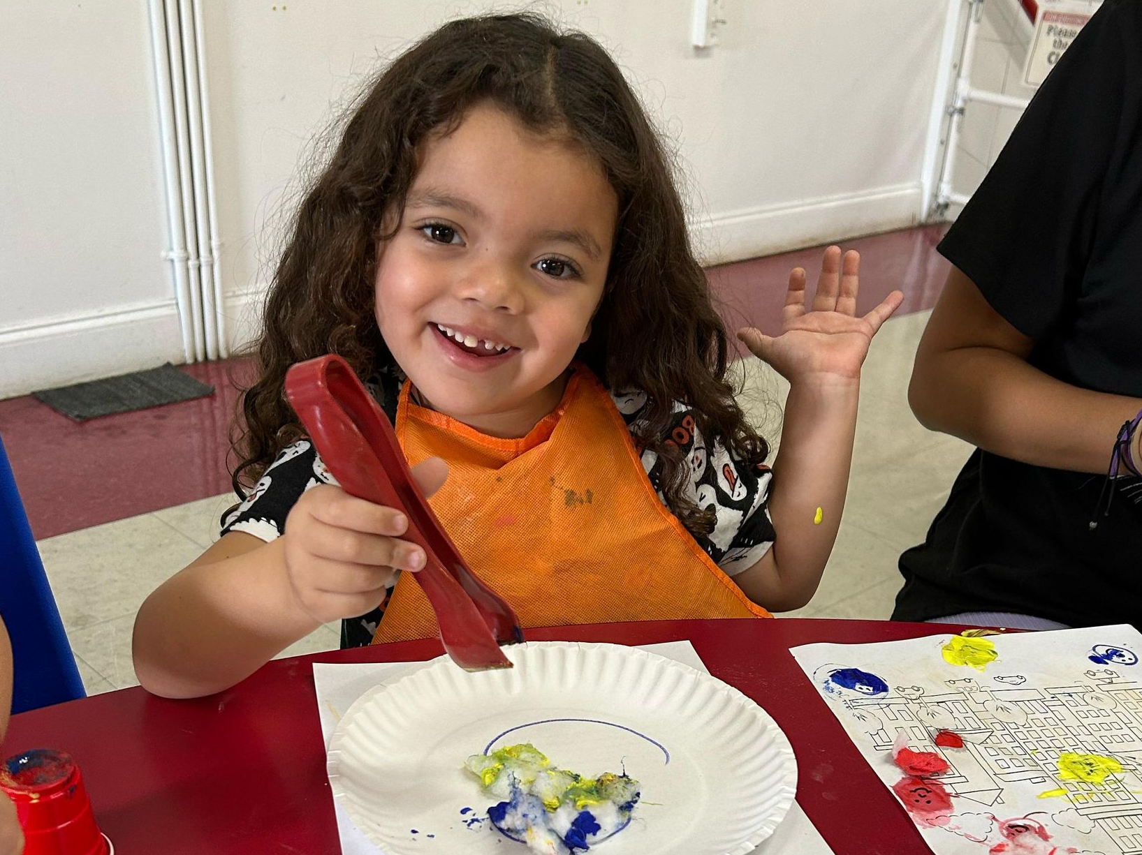 Smiling child holding a red paintbrush at a table with paper and paint in a classroom