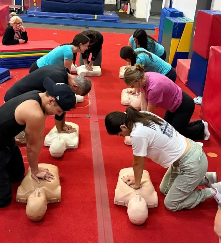 Group practicing CPR on training mannequins in a gym, with red mats and colorful equipment.