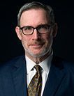 Man in glasses, suit, and tie against a dark background, looking forward.
