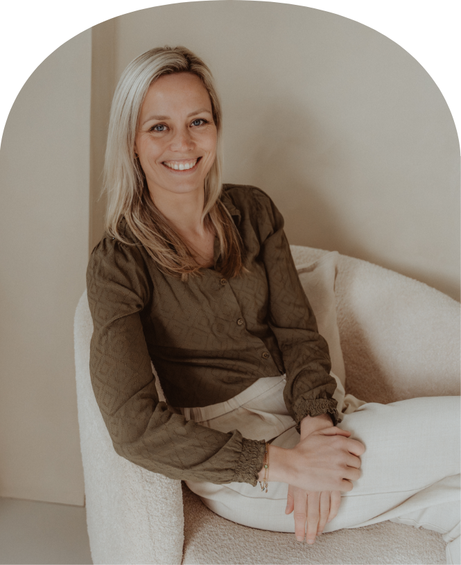 Smiling blonde woman sitting on a cream chair in a beige room, wearing a brown top and white pants
