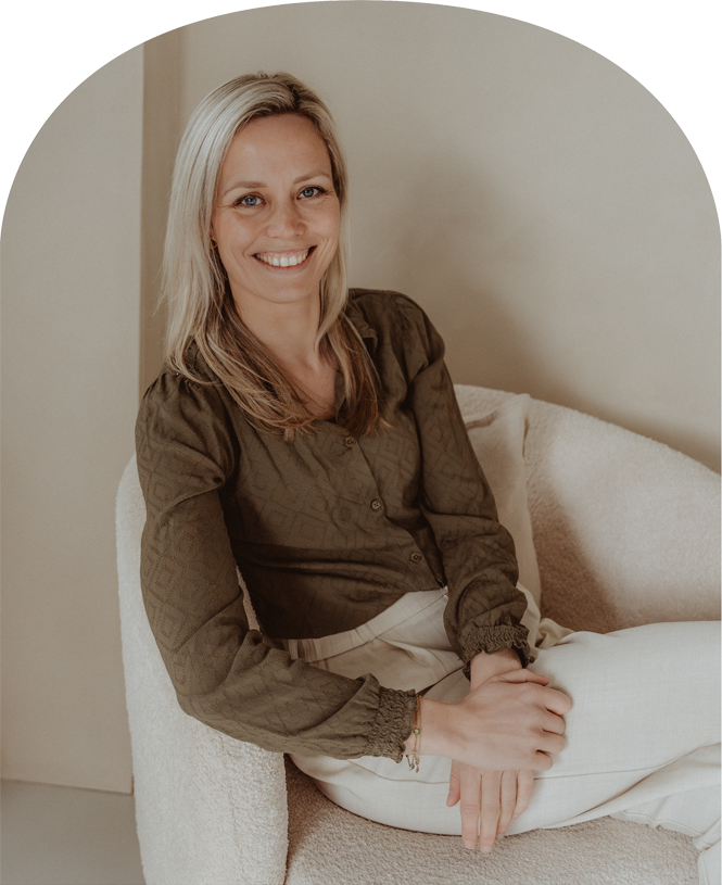 Smiling blonde woman sitting on a cream chair in a beige room, wearing a brown top and white pants
