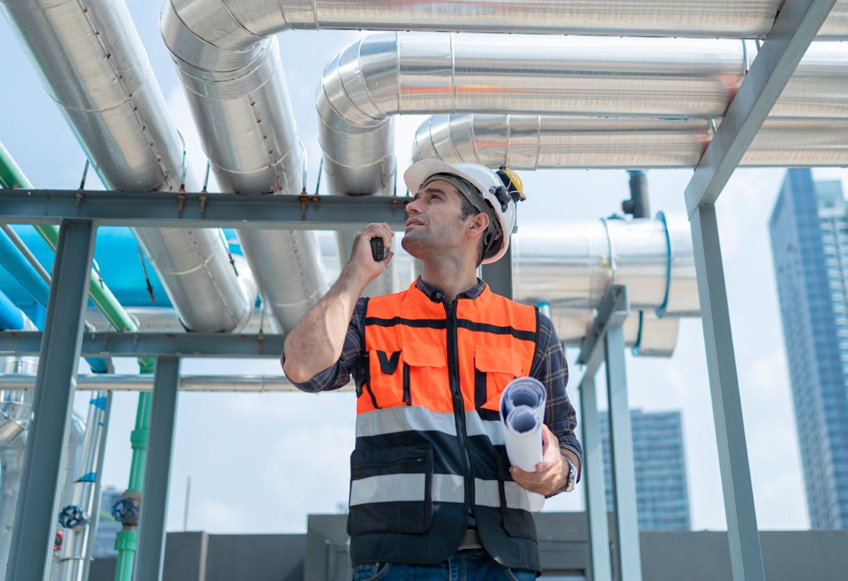 Construction worker in orange vest and hard hat inspecting pipes, using a radio, outdoors.