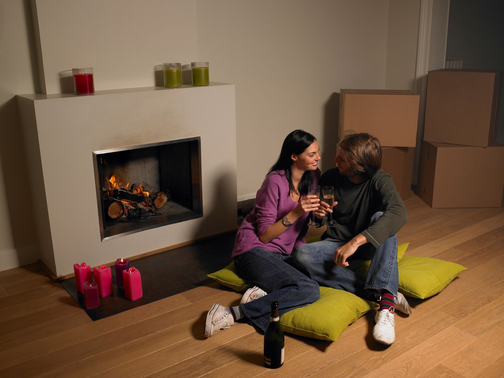 Couple toasting near fireplace in a new home, sitting on pillows, surrounded by boxes.