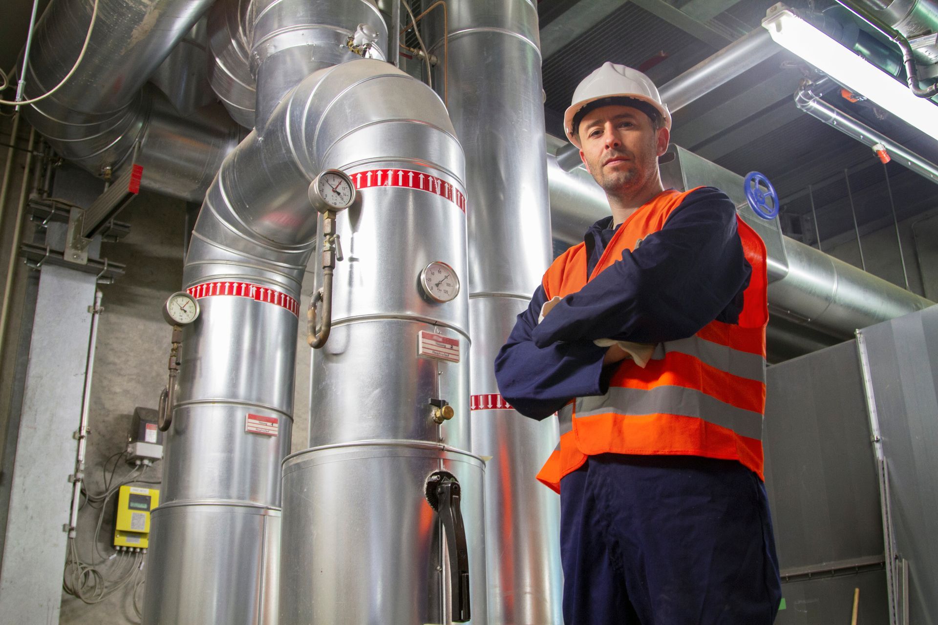 Industrial worker in orange safety vest stands beside large metal pipes in a factory room