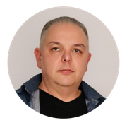 Retrato de um homem com expressão neutra, vestindo camisa escura e jaqueta jeans, sobre um fundo claro.