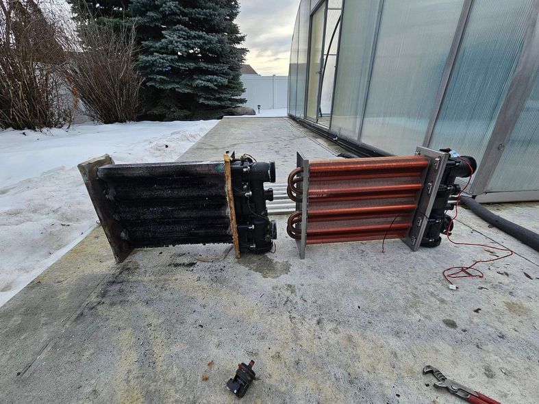 Two heat exchangers, one black and one copper, on a concrete surface with snow in the background.